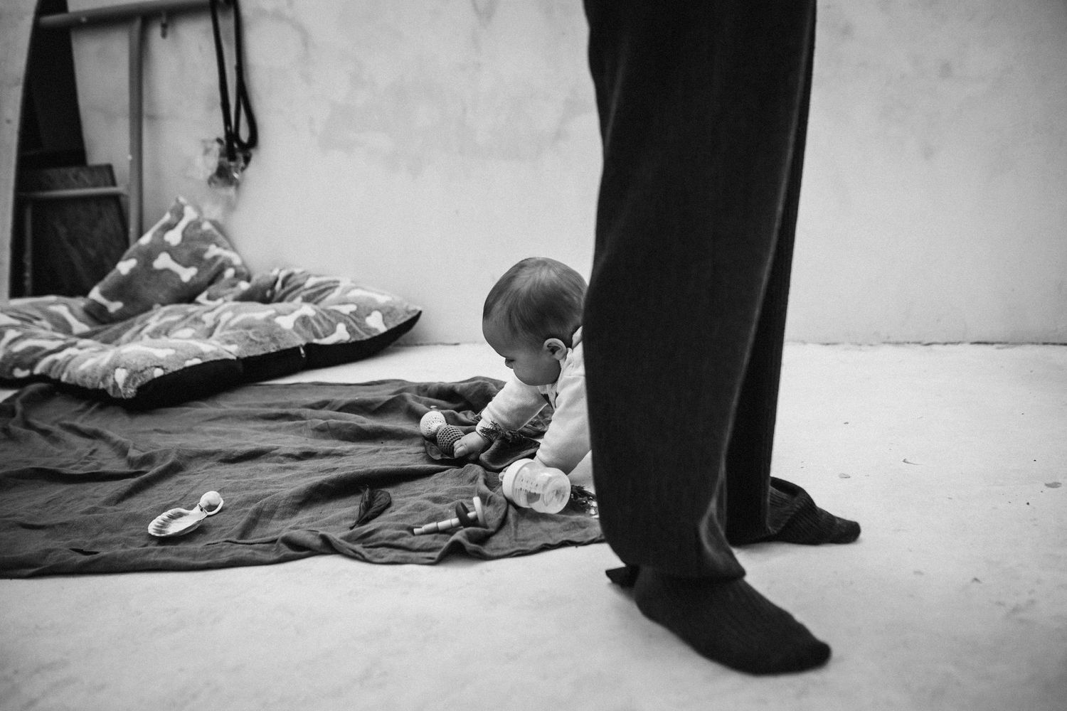 A small child lies on a blanket on the floor while adult legs stand nearby in black and white photograph.