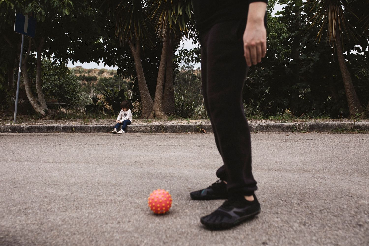Person standing on a court with an orange basketball while another person sits in the background among trees.