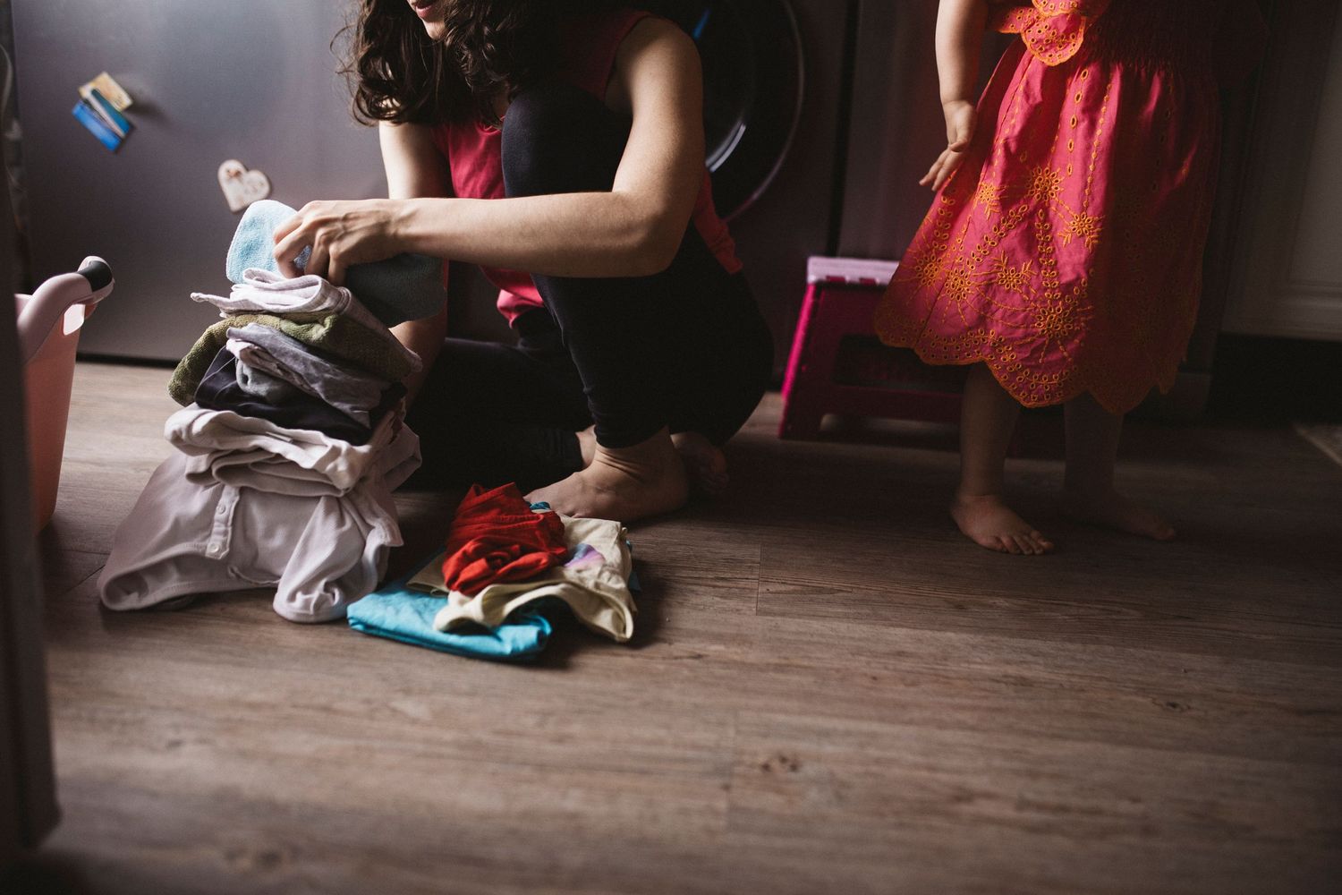 Pile of folded laundry sits on wooden floor while someone in a red dress walks by in the background.