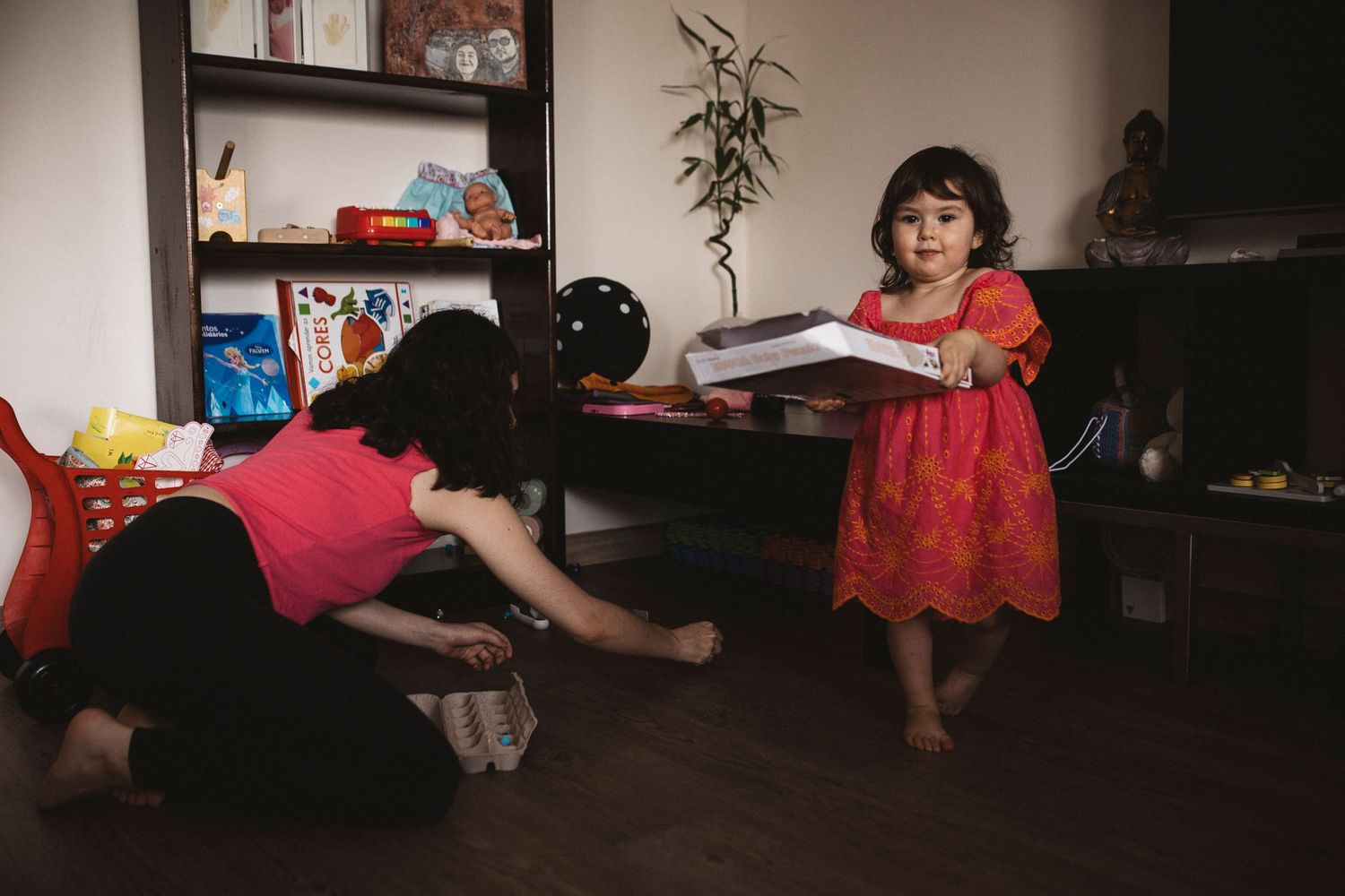 A child in a red dress stands in a dimly lit living room while someone cleans the floor.