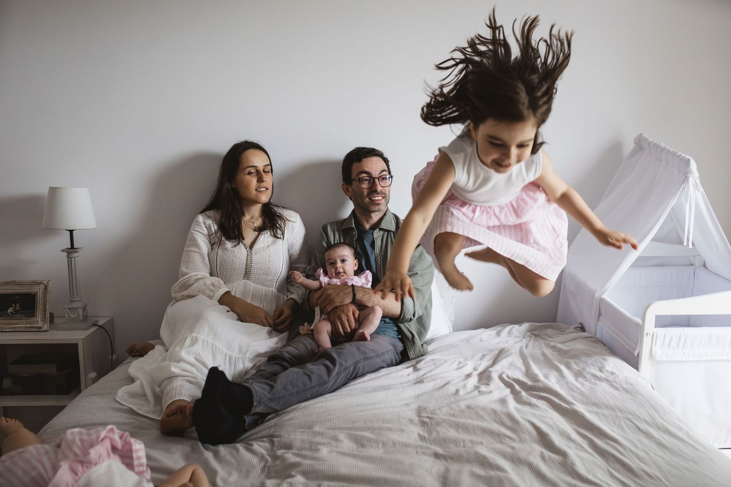 A family plays and jumps together on a white bed in a bright bedroom.