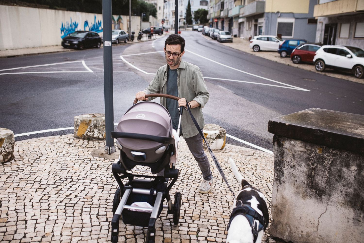 A person walks with a baby stroller along a cobblestone street in an urban neighborhood with parked cars.
