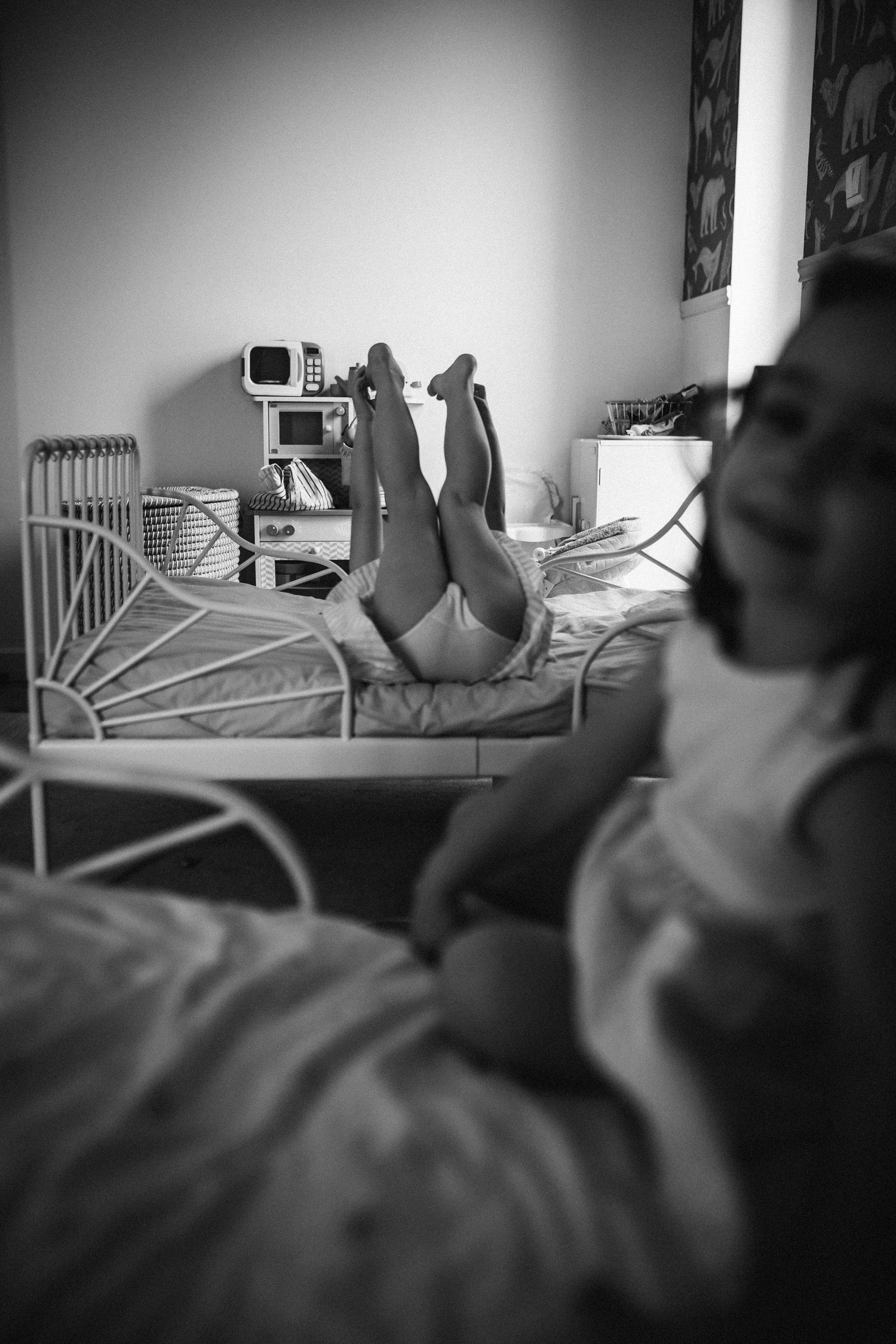 Black and white photo showing feet propped up on a bed in a cozy bedroom with a radiator and bookshelves in view.