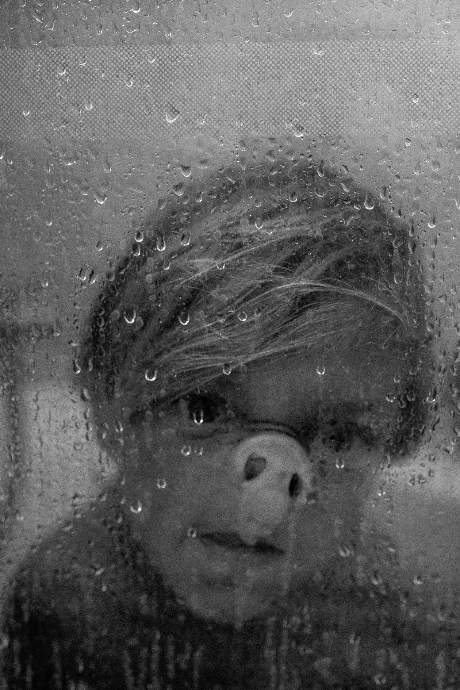 A black and white photograph taken through a rain-covered window shows a distorted figure behind steamy glass.