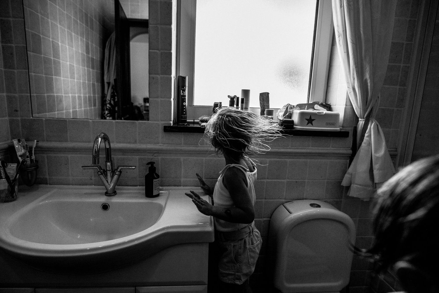 Black and white photo of a small child reaching up to a bathroom sink while standing near a toilet.