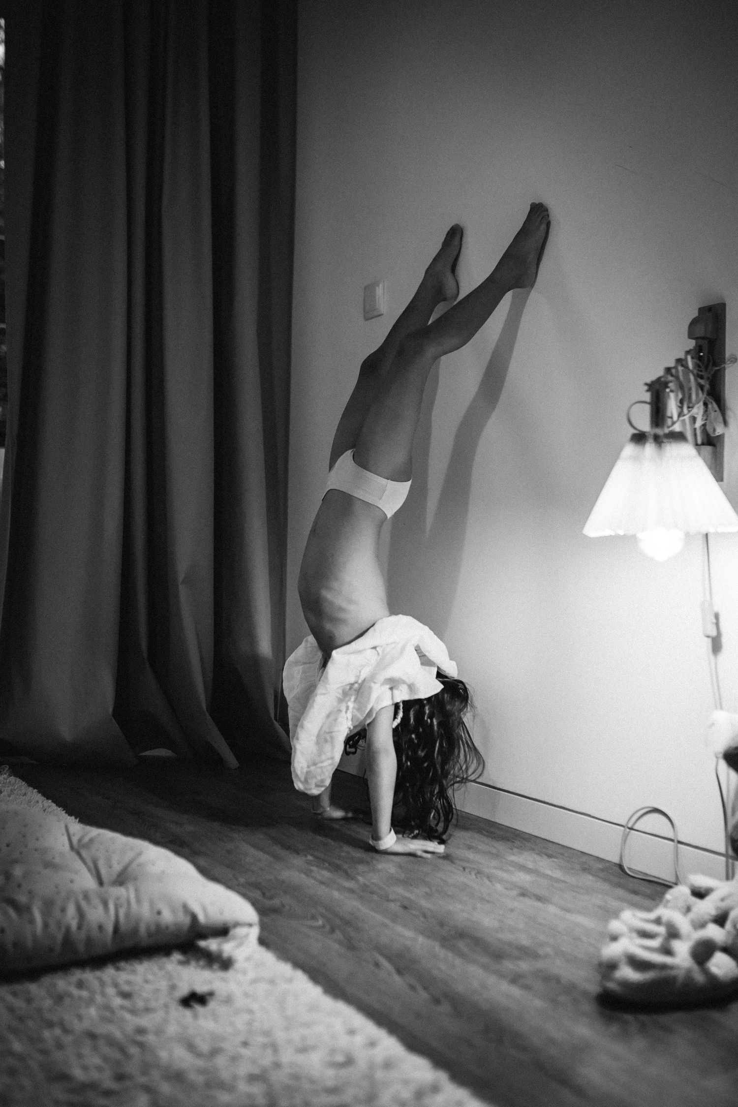 Black and white photo showing someone doing a handstand against a wall near a lit lamp and curtains in a bedroom.