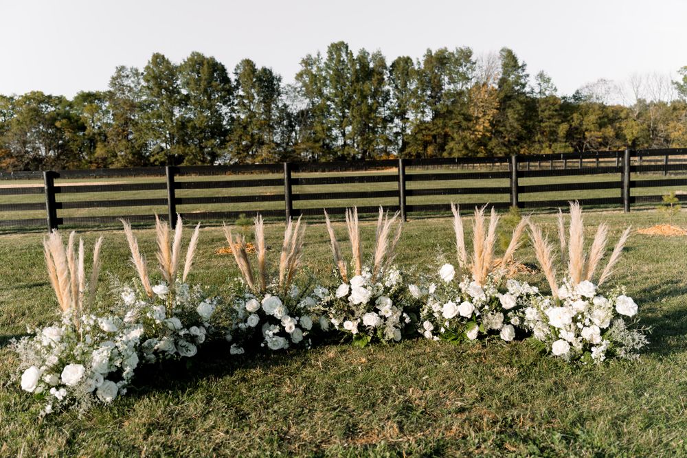 Low floral arrangement with white flowers and pampas grass decorates a rustic outdoor venue with wooden fence.