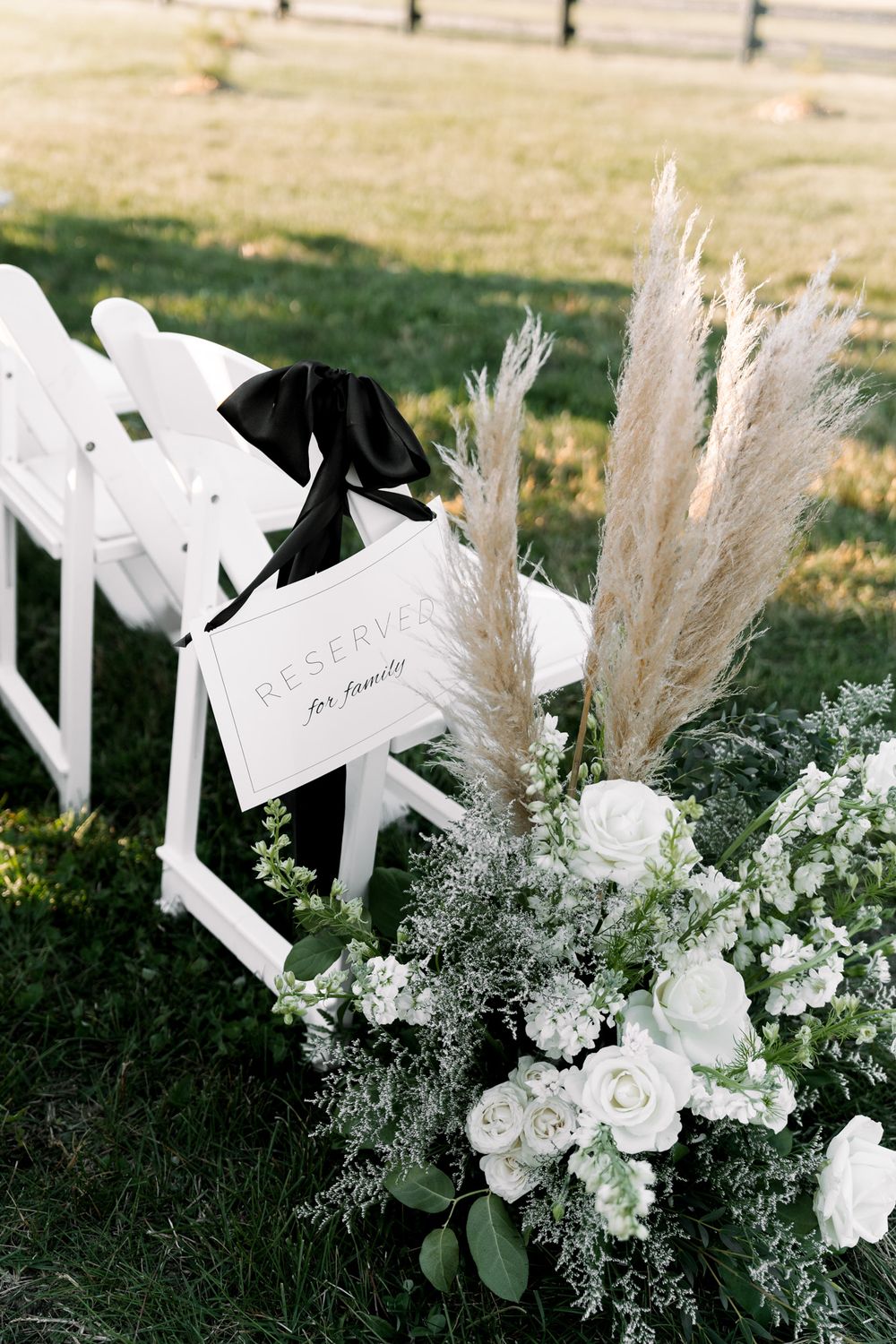 White folding chairs and elegant floral arrangements with pampas grass decorate an outdoor wedding ceremony setup.