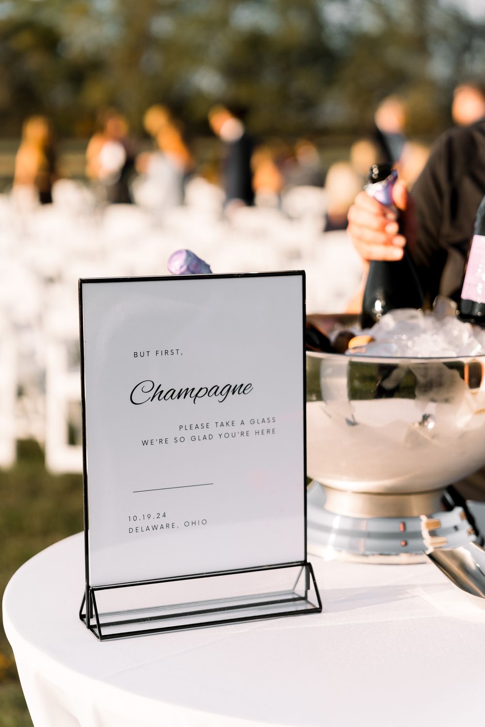 Elegant champagne bar sign displayed in black frame on white table with blurred glasses and decor in background.