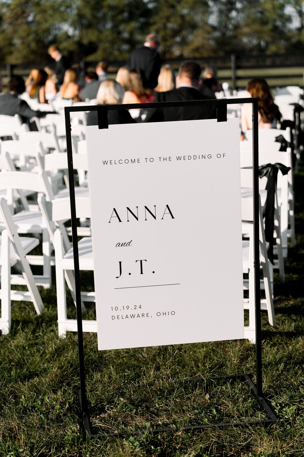 A wedding welcome sign with clean typography displaying 'Anna and J.T.' on a black stand with white chairs in the background.