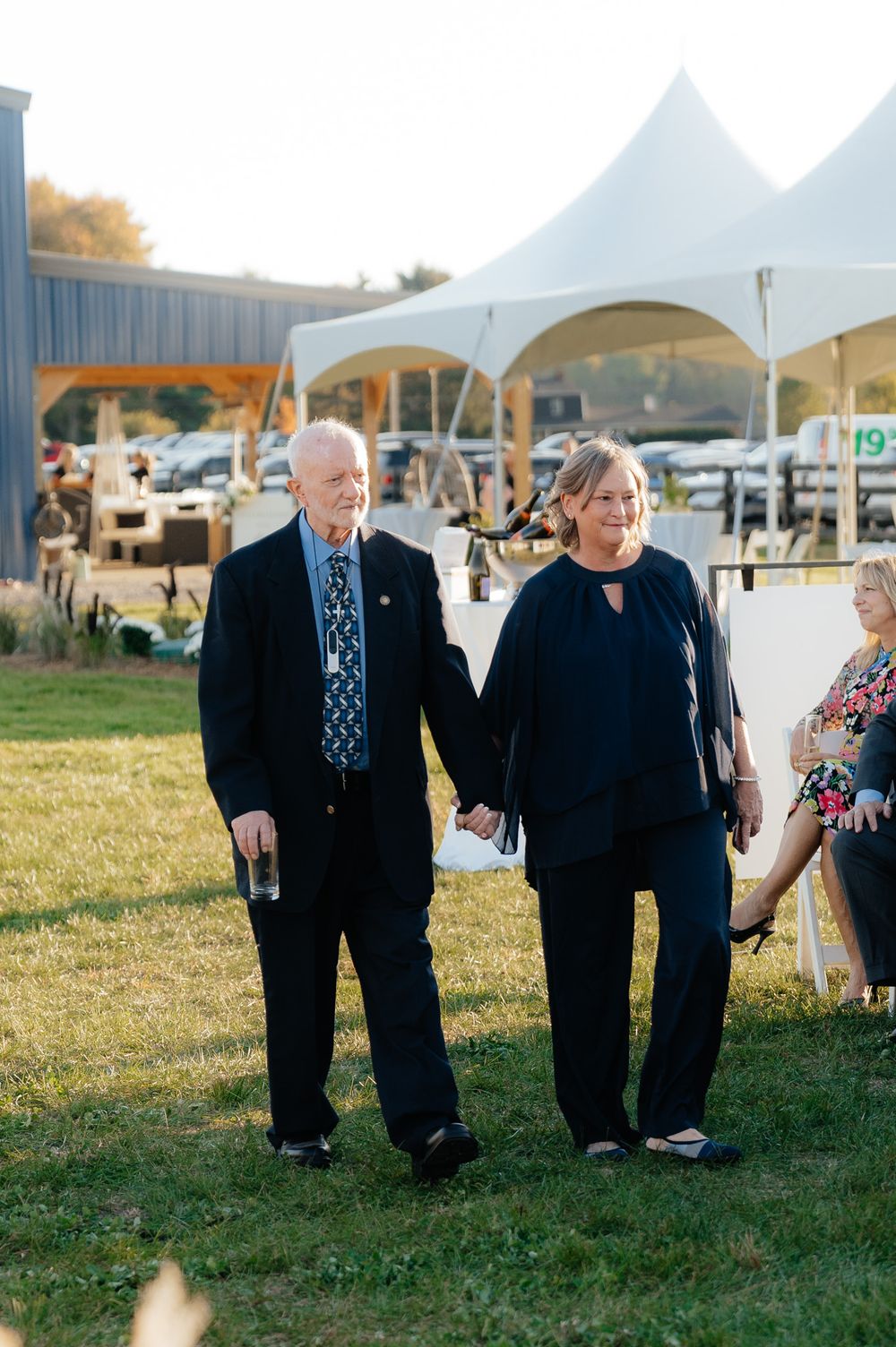 Two people holding hands while walking on grass at an outdoor event with white tents in the background.