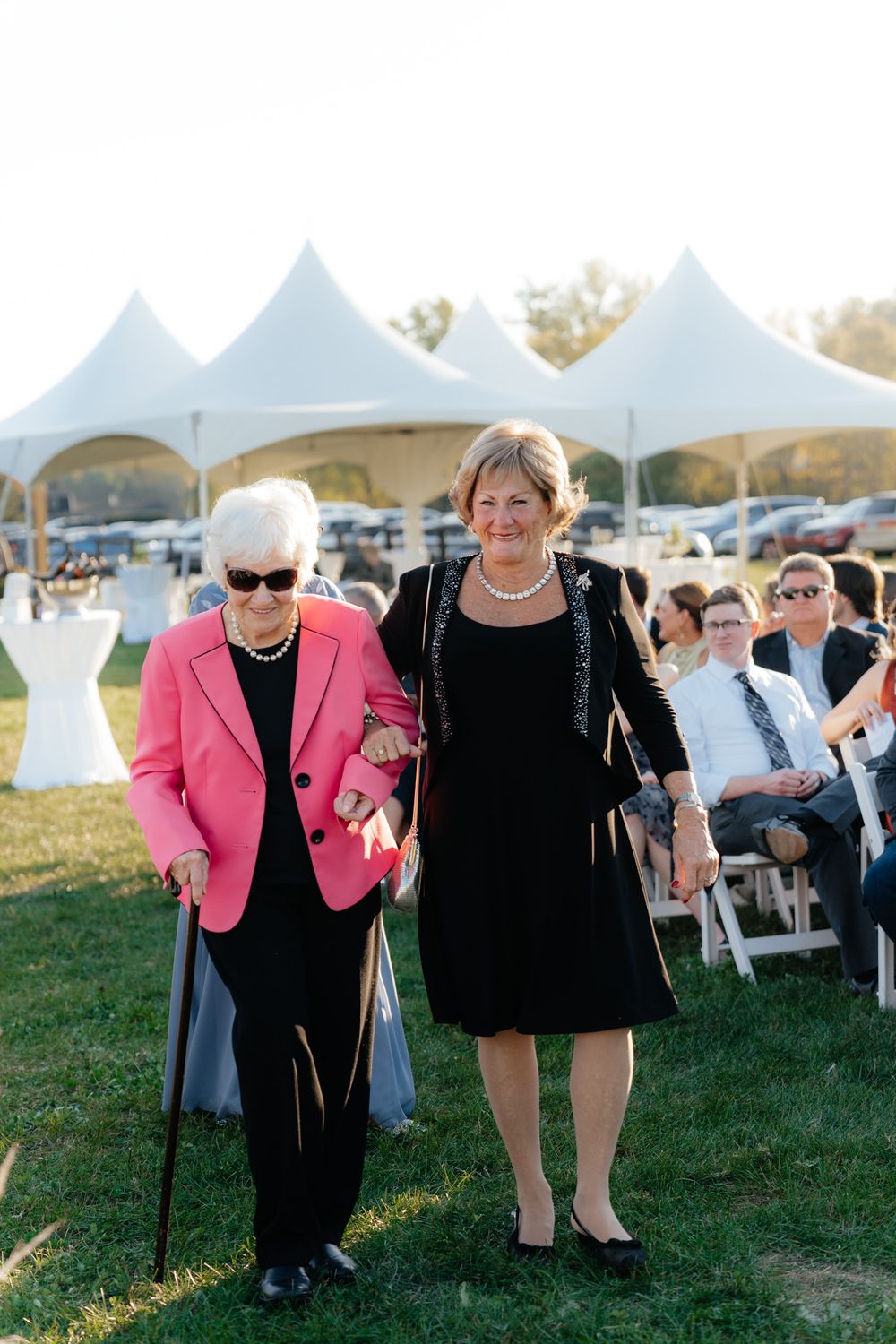 Two guests at an elegant outdoor wedding with white tents, one wearing a bright pink blazer and using a cane.