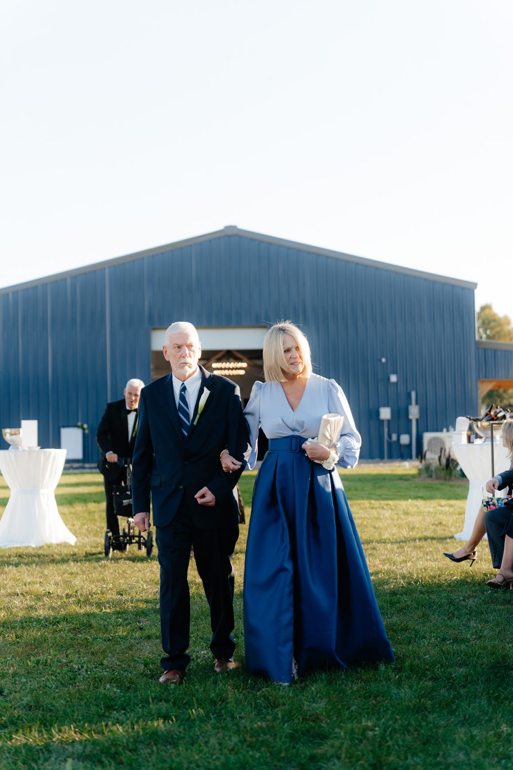 Outdoor wedding scene featuring guests in formal attire near a blue metal barn structure.