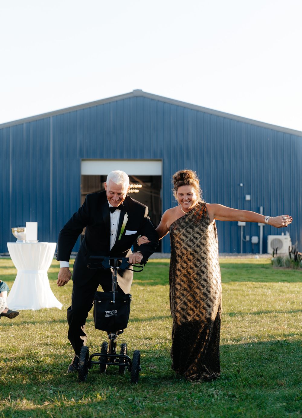 A joyful moment outside a blue barn as two people share a laugh while walking together at a wedding celebration.