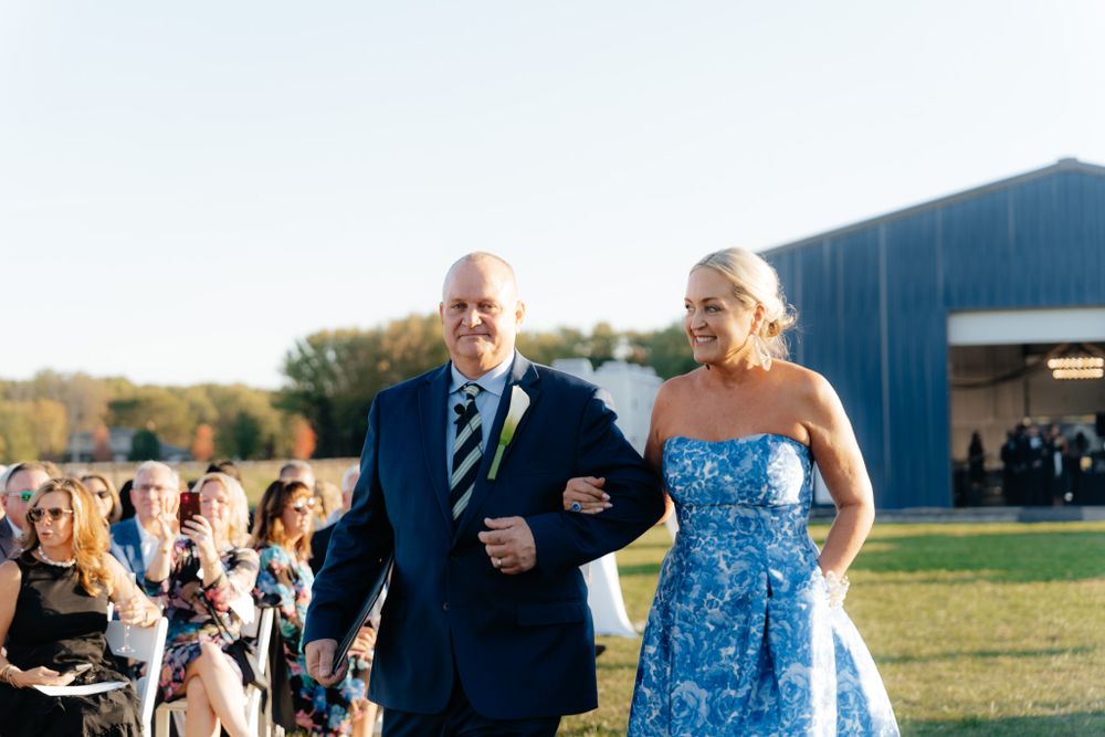 A wedding ceremony processional takes place outdoors with attendees seated on white chairs on a sunny day.
