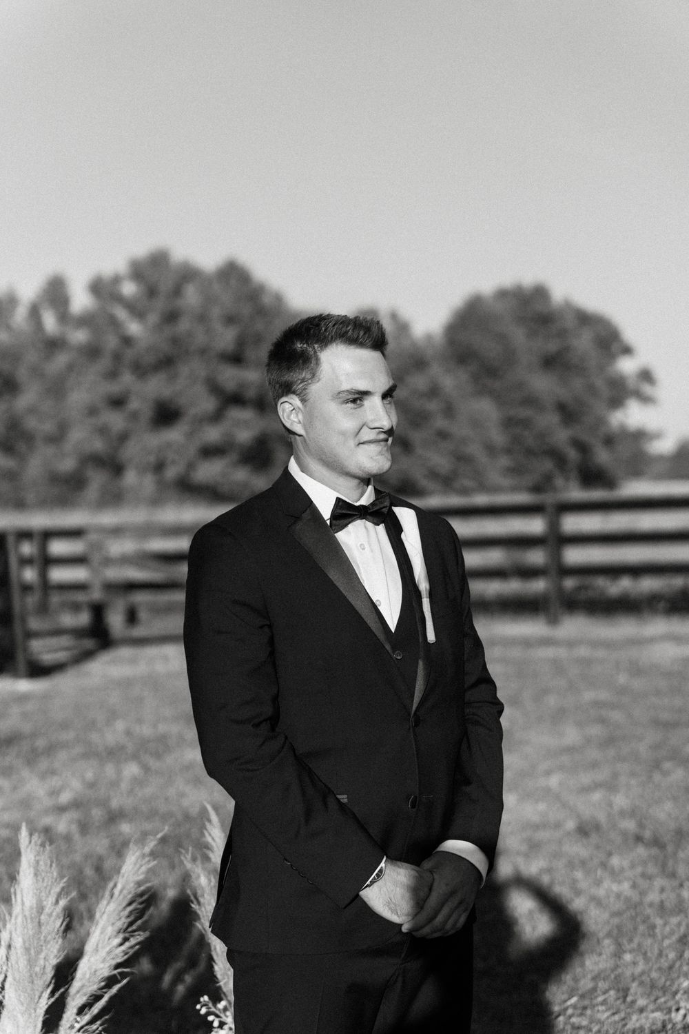 Black and white photo of groom in formal tuxedo standing outdoors at a ranch with wooden fence in background.