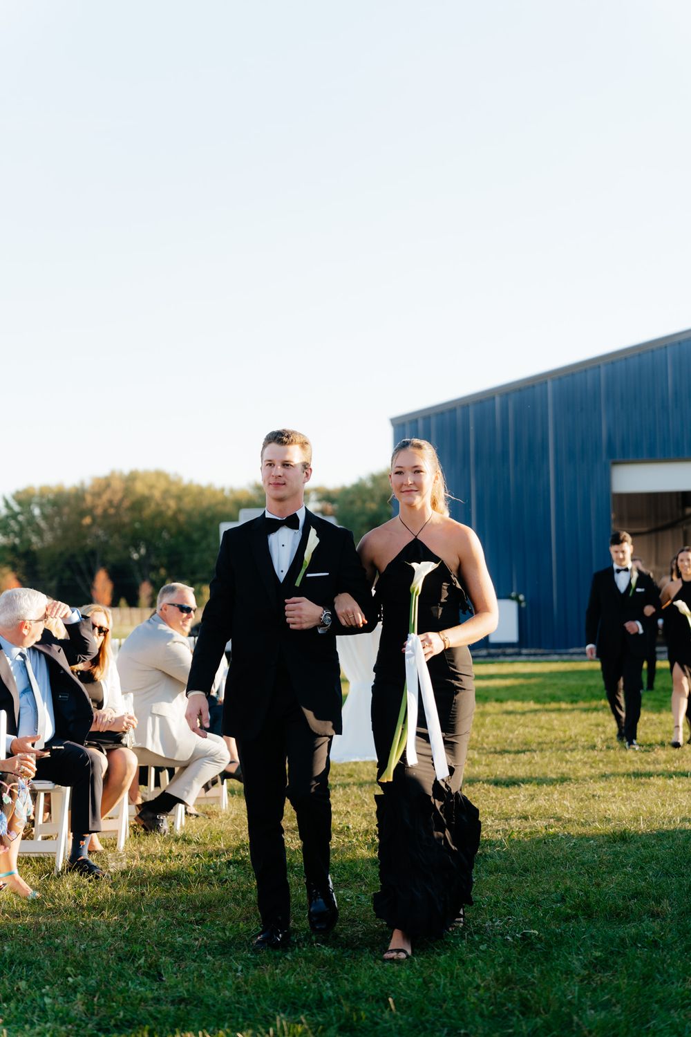 Wedding attendants walking on a grassy lawn near a blue barn building during a sunny day.