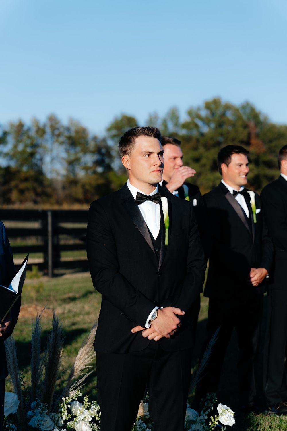 Groomsmen in black tuxedos standing together during an outdoor wedding ceremony.