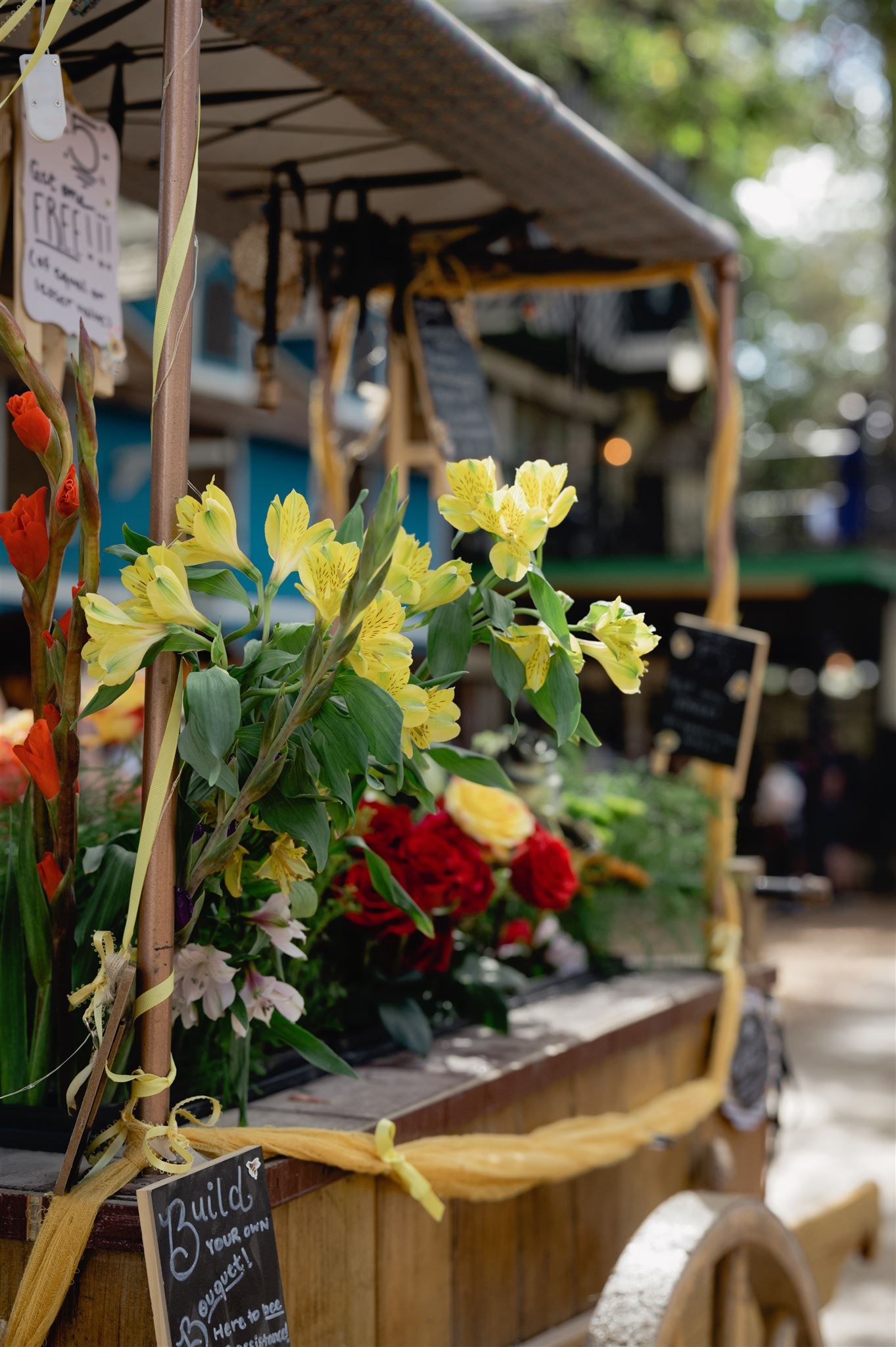 Wooden market stalls displaying bright yellow daffodils and red flowers with sunlight filtering through trees.
