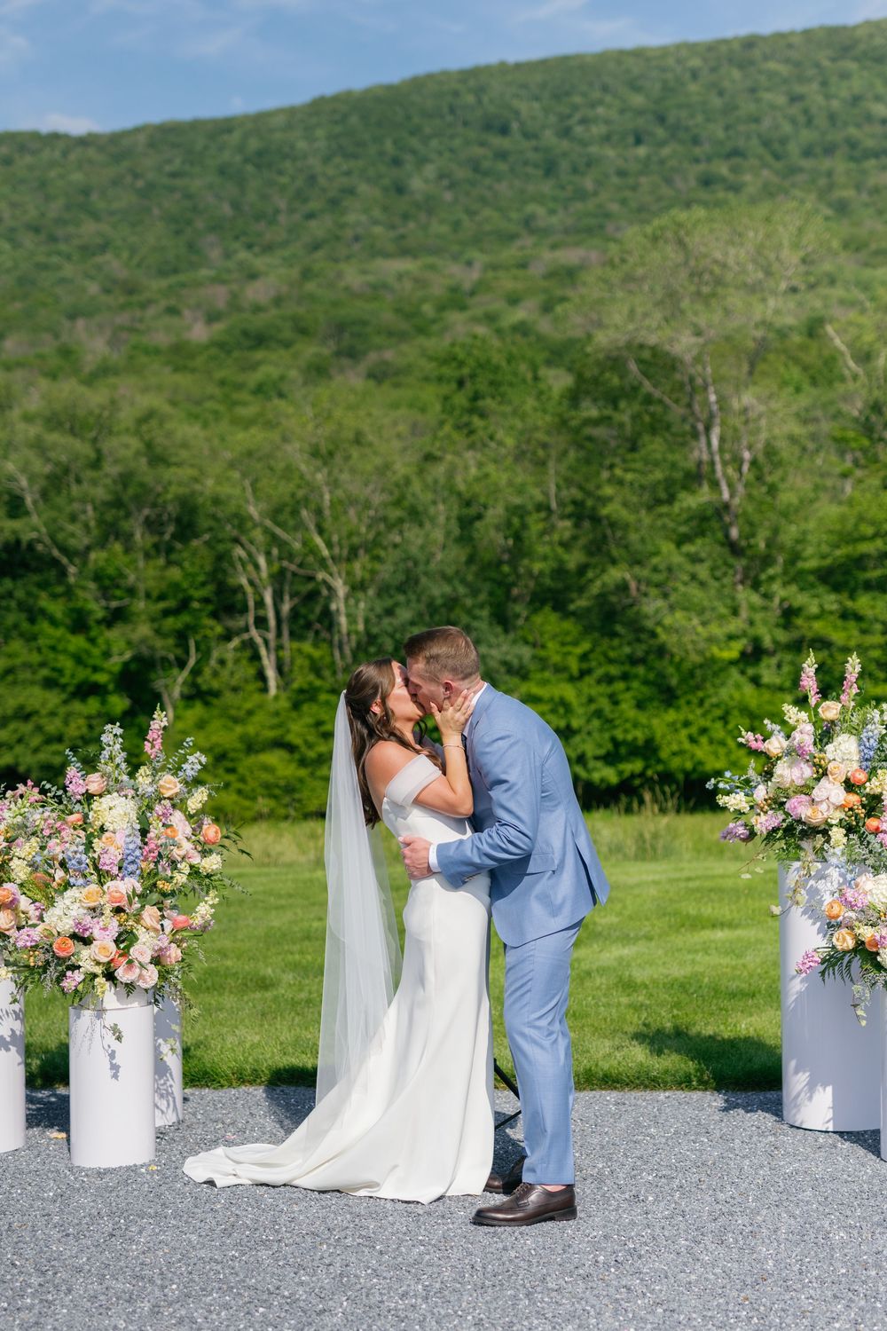 A couple shares their first kiss during an outdoor wedding ceremony with pink floral arrangements and mountain backdrop.