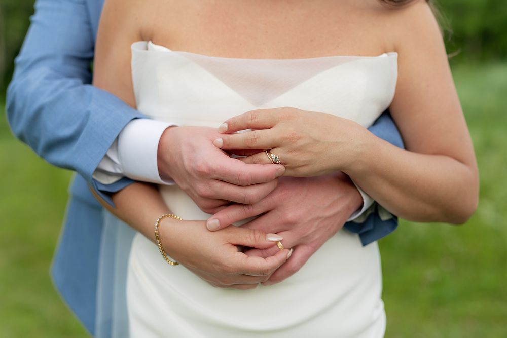 Close-up of newlywed couple's hands embracing, with white wedding dress and blue suit visible in outdoor setting.