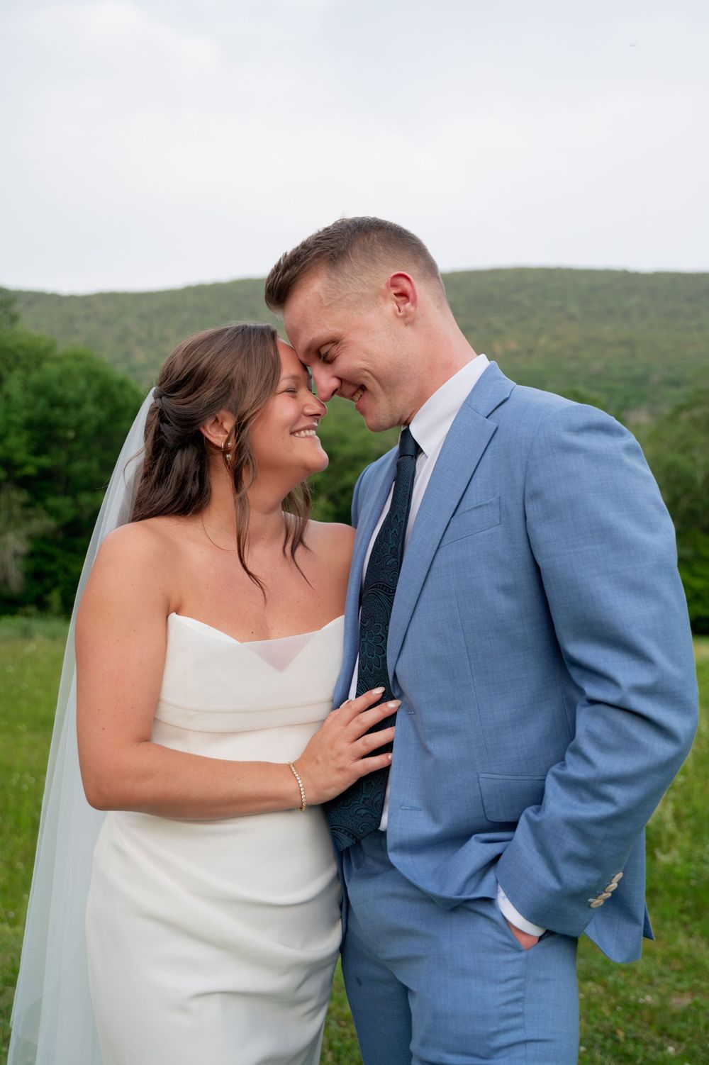 A couple in wedding attire share an intimate moment against a scenic green hillside backdrop.