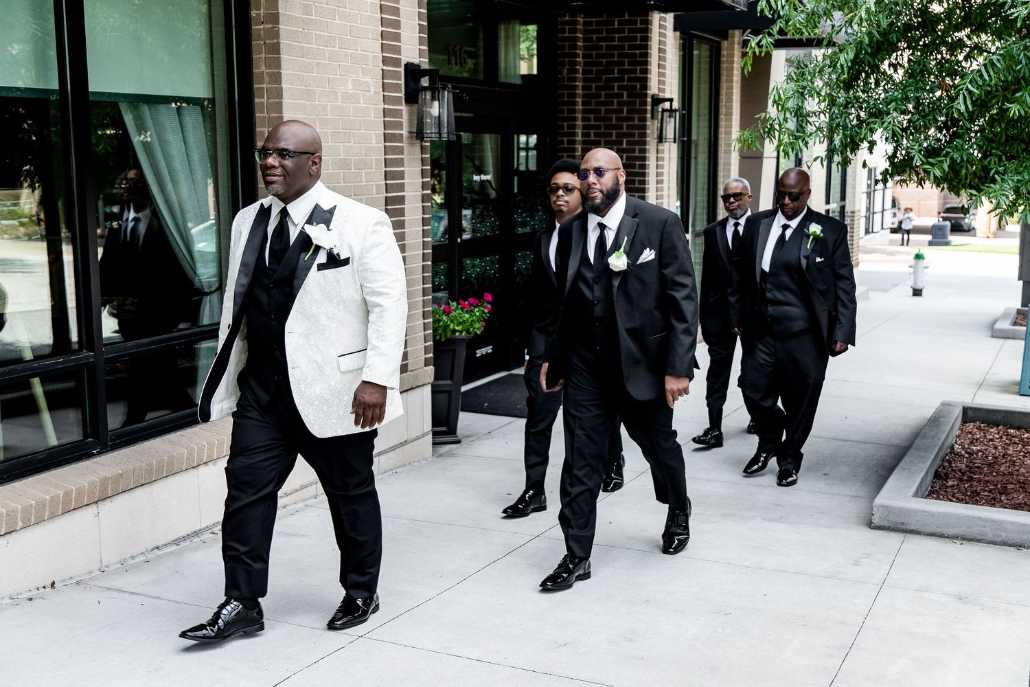 Groomsmen in black and white formalwear walk together down a sunny urban sidewalk outside a brick building.