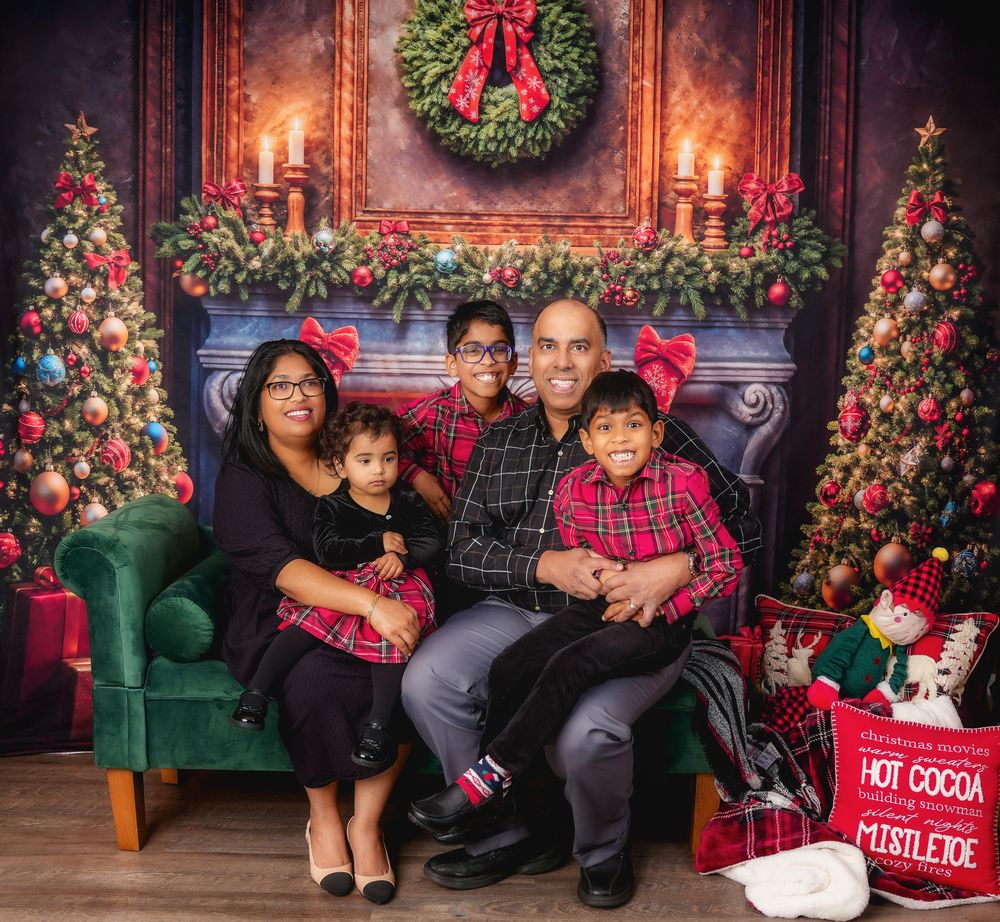 A cozy family Christmas portrait with decorated trees, a fireplace mantel, wreath, and wrapped presents by a green velvet couch.