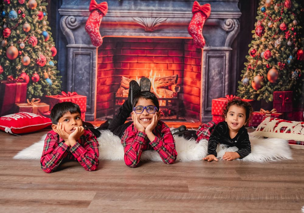 Children in matching plaid pajamas lying on fur rug in front of decorated Christmas fireplace with stockings.