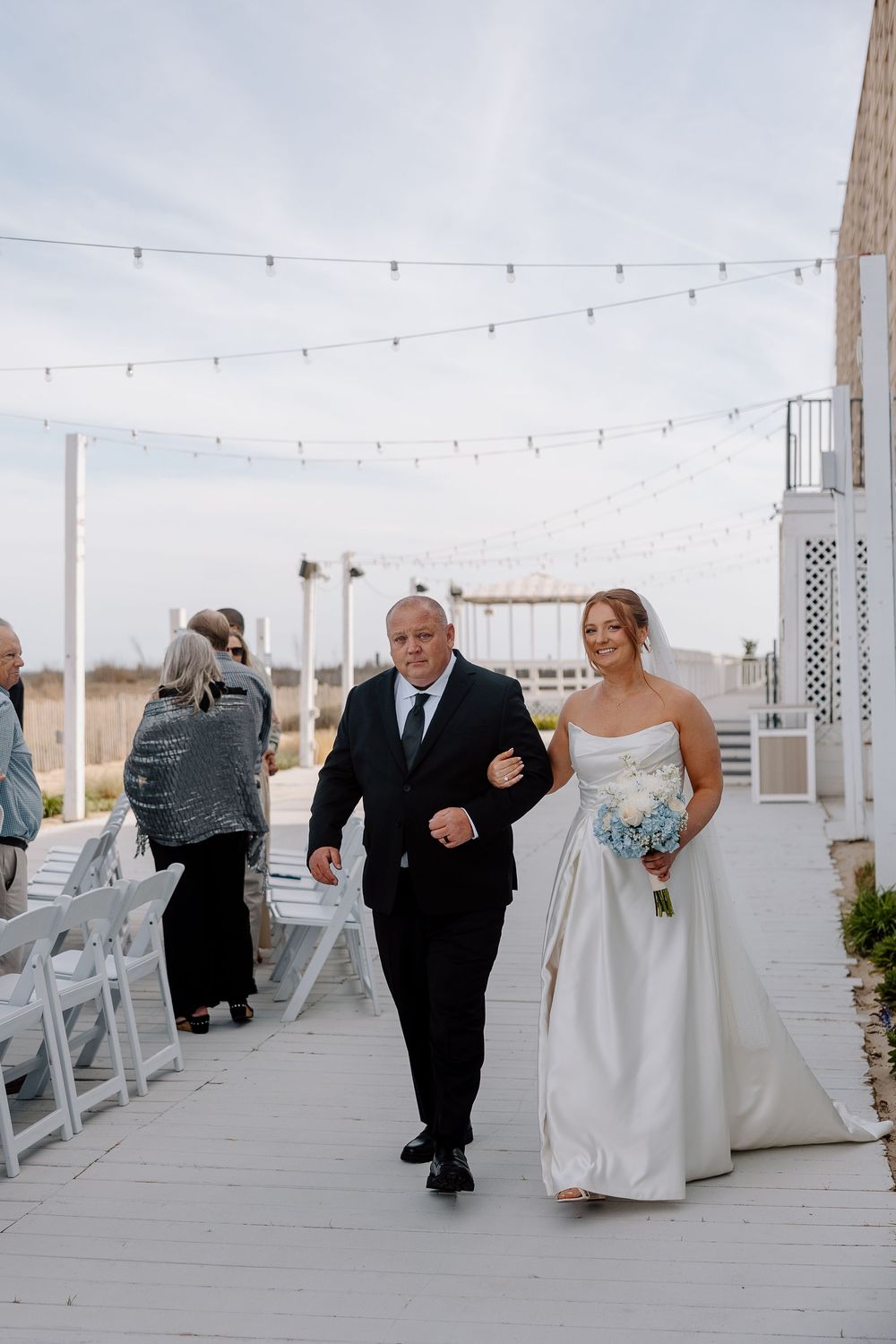 Four sequential photos show wedding party members walking down a white boardwalk decorated with string lights at dusk.