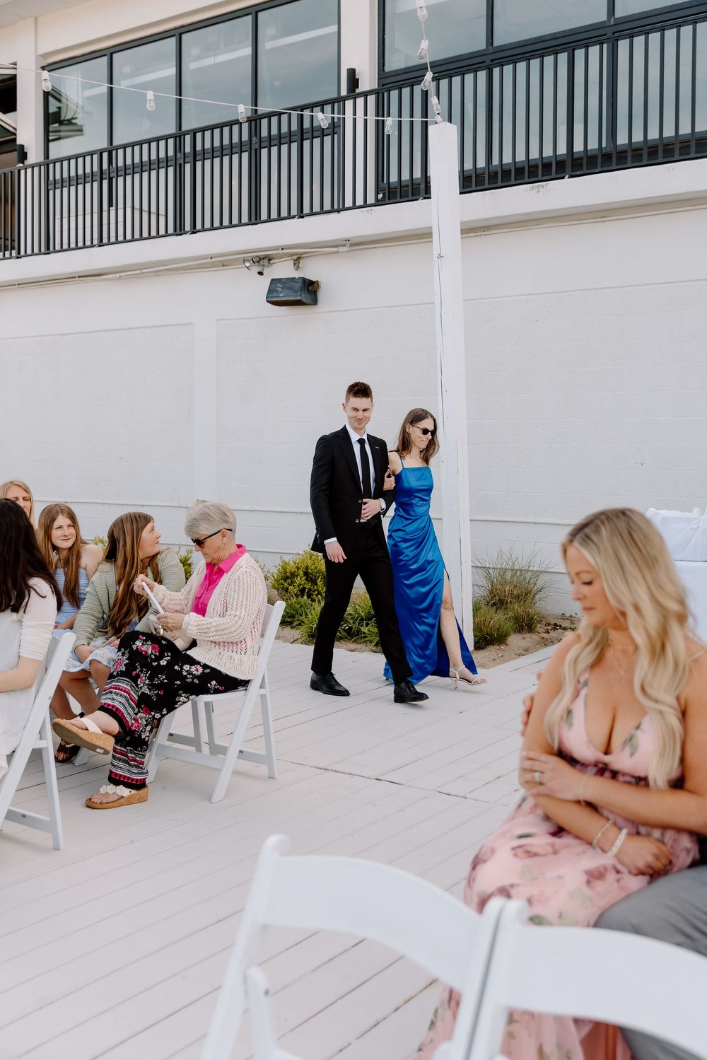 Guests in formal attire gather at an outdoor venue with white chairs and balcony railing for an elegant event.