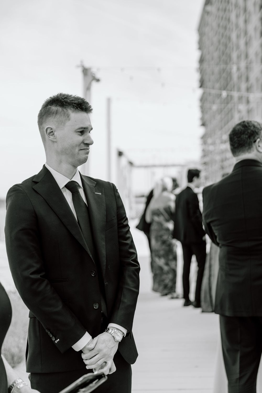 Black and white photo of someone in a formal suit waiting outdoors with others in an urban setting.