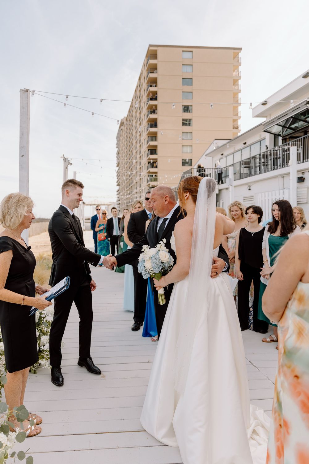 A group in formal attire gathers on an outdoor terrace with tall apartment buildings visible in the background.