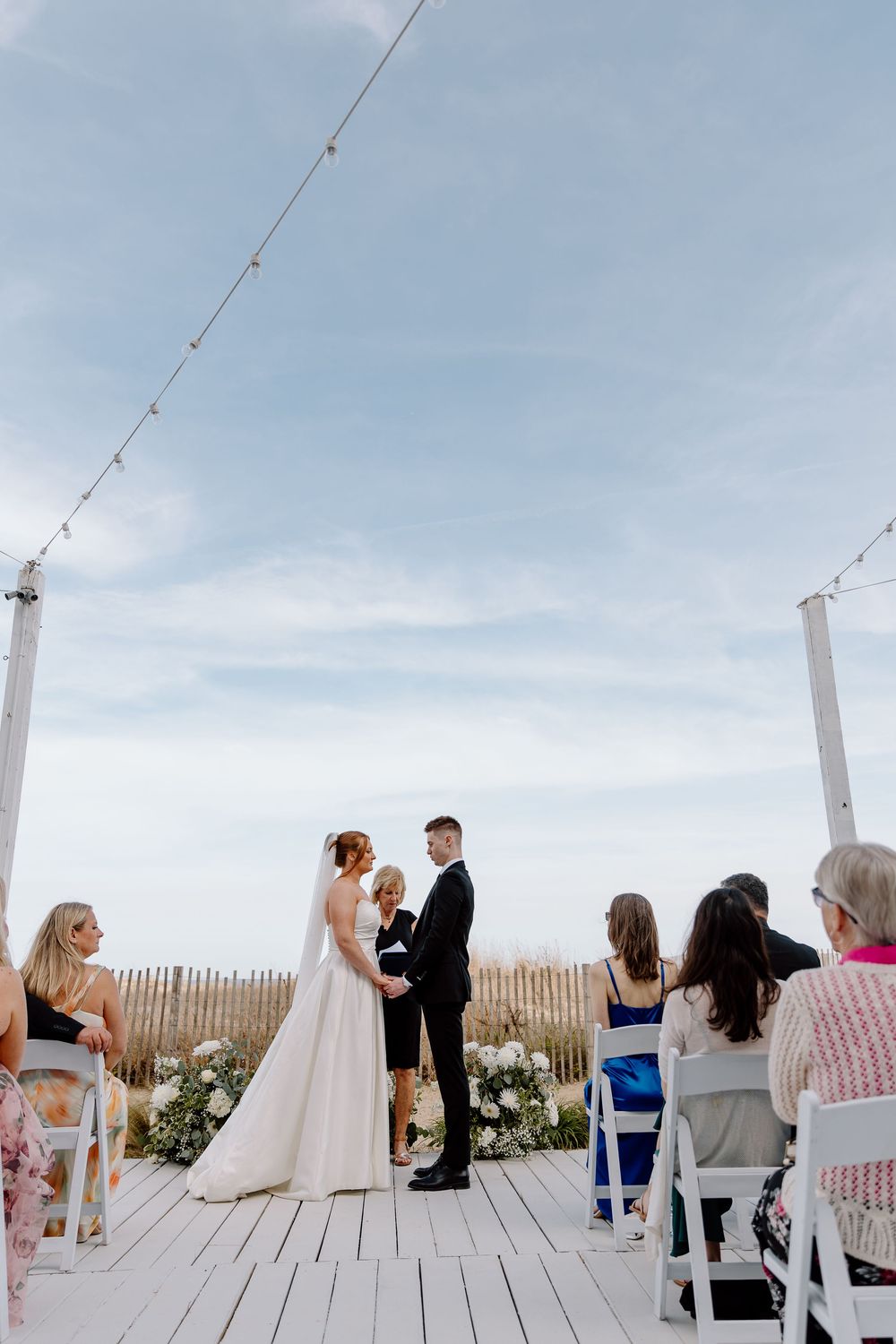 An outdoor beach wedding ceremony takes place on a wooden deck with string lights overhead and guests seated on white chairs.