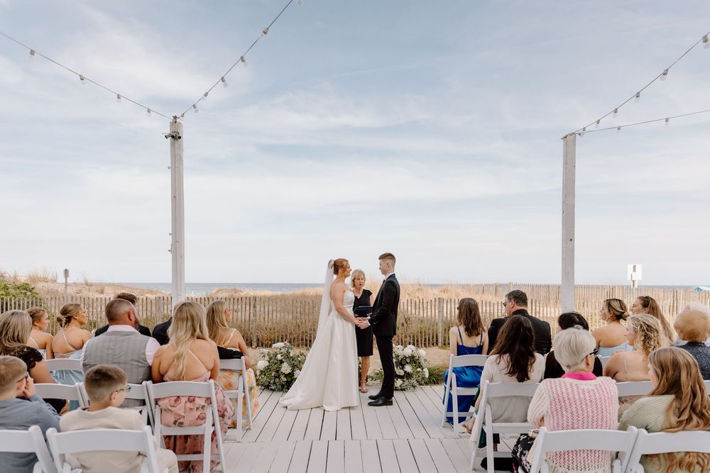 An intimate beach wedding ceremony takes place on a wooden deck with string lights and ocean views in the background.
