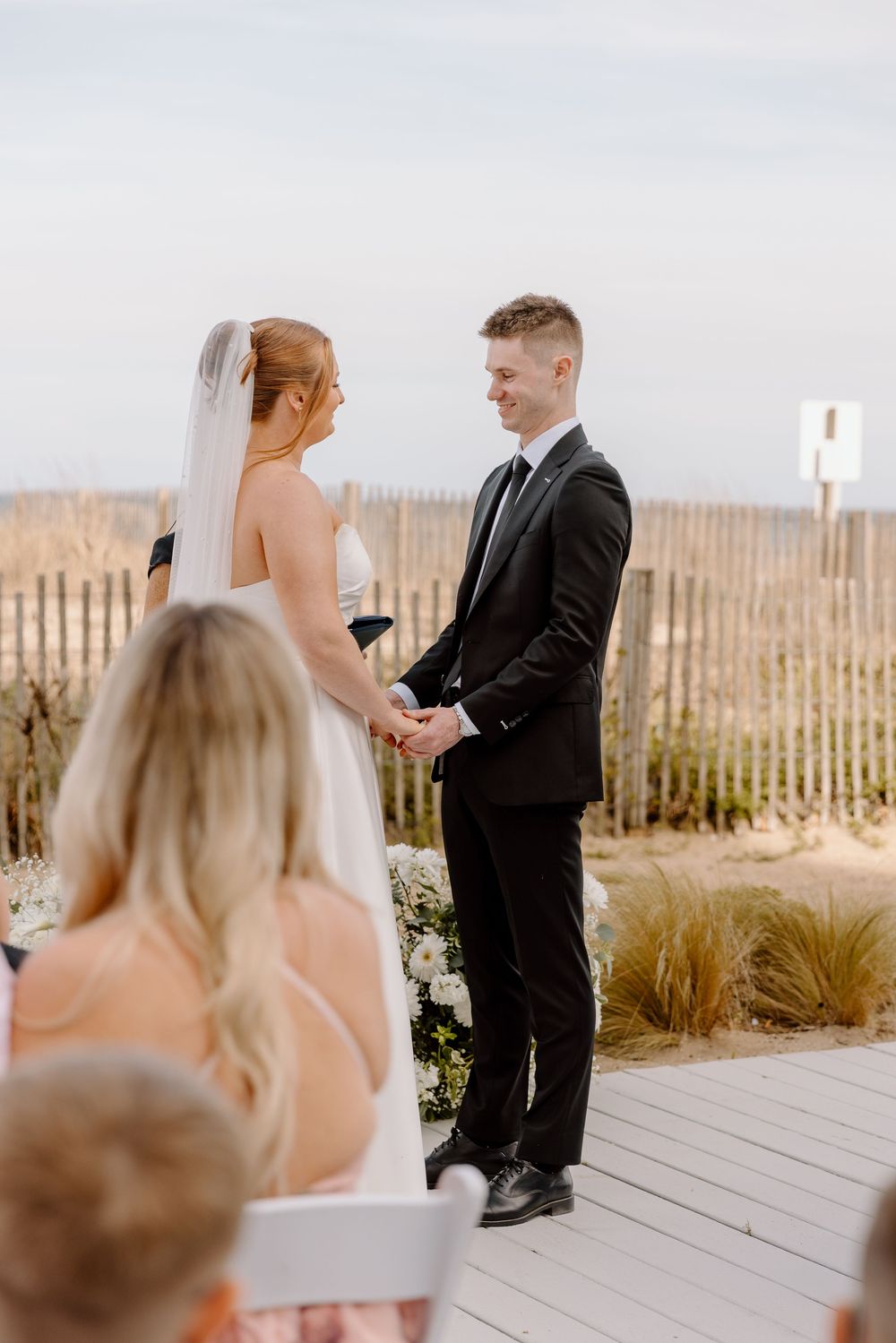 A romantic seaside wedding ceremony where a couple stands on a wooden boardwalk with beach fencing and dunes in the background.