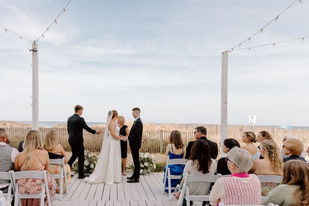 A scenic beachside wedding ceremony takes place on a wooden deck with string lights and ocean views in the background.