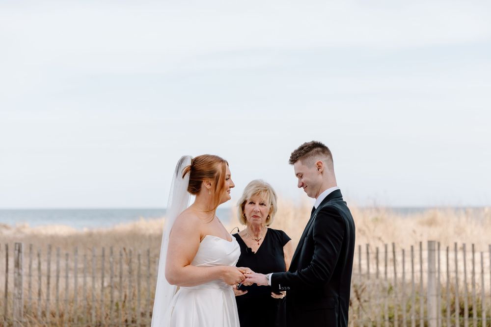 A couple exchanges wedding vows on a scenic beach with wooden fences and dunes in the background.