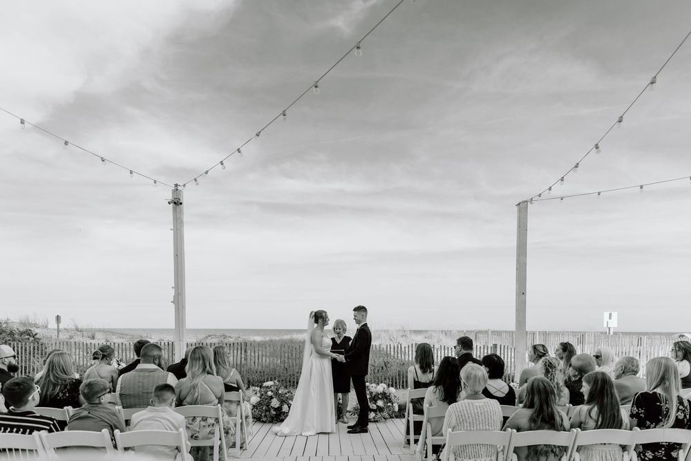 Black and white photograph of a seaside wedding ceremony on a deck with string lights and ocean views.