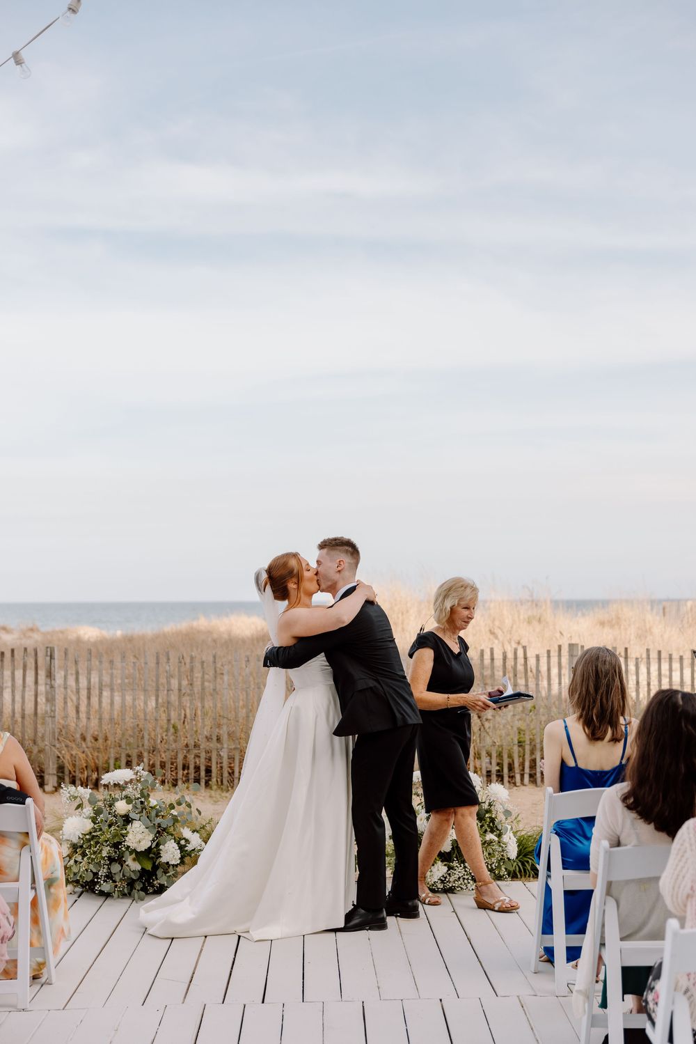 Wedding guests observe an intimate beachside ceremony from white chairs on a wooden deck with coastal scenery.