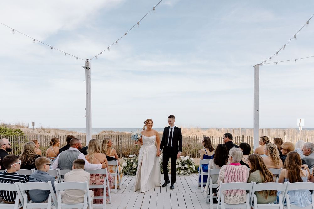 Beautiful outdoor wedding ceremony on a wooden deck overlooking sandy dunes with string lights and white chairs.