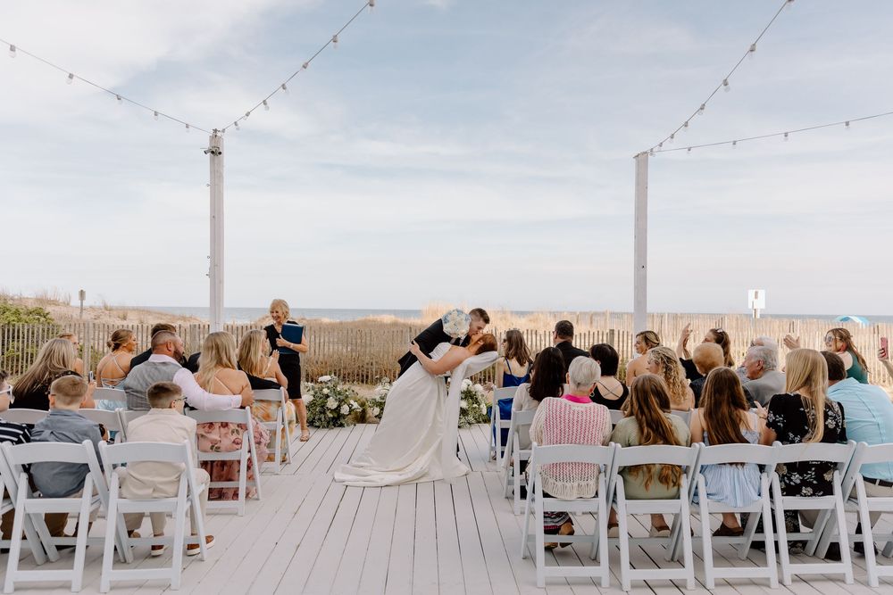Panoramic view of guests seated in white chairs on a wooden deck with string lights during an outdoor seaside event.