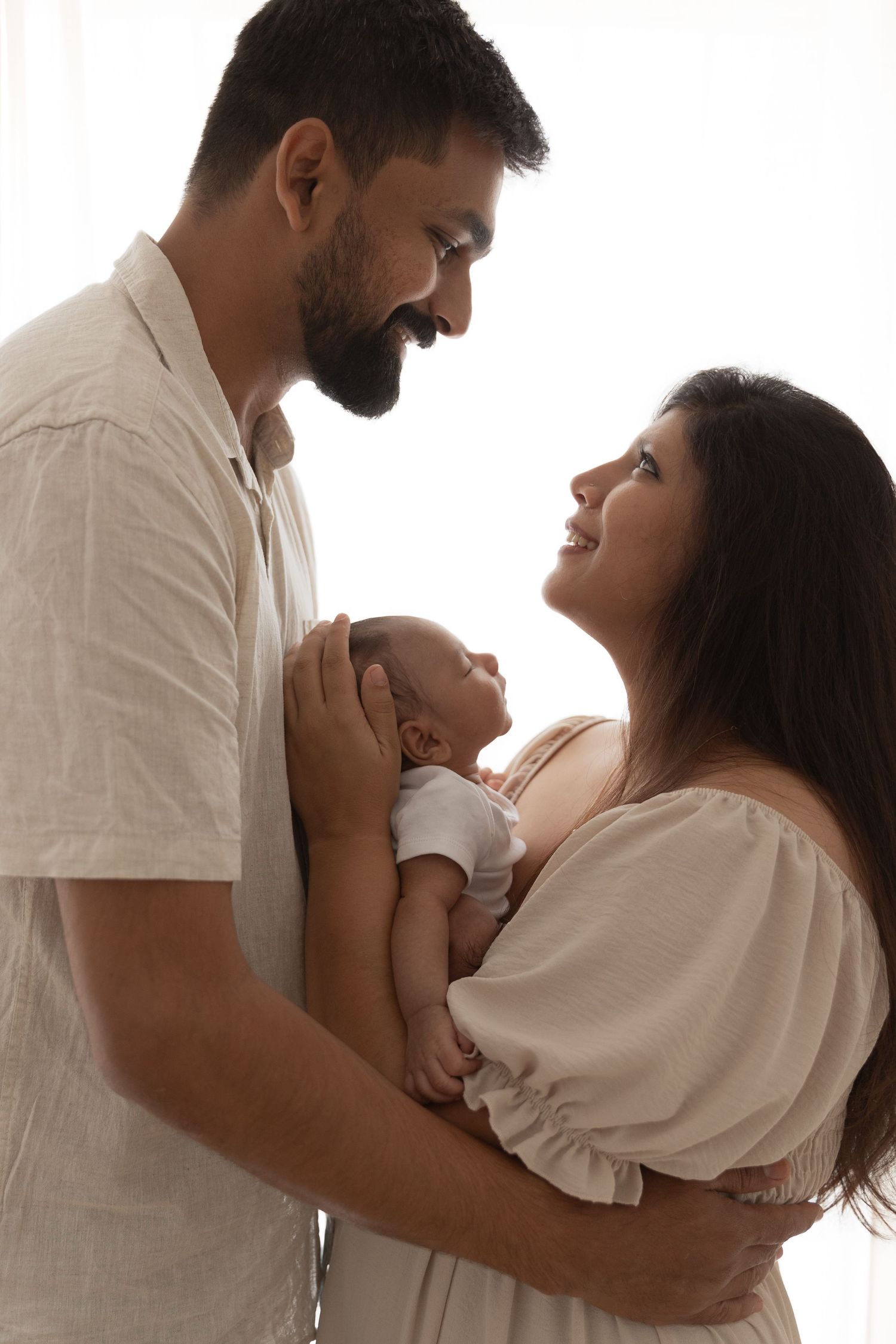 Loving couple in white outfits tenderly embracing their newborn baby in a bright studio portrait.