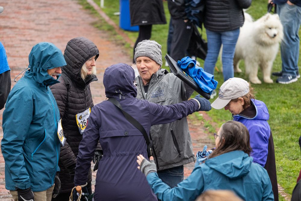 Group of people in winter coats gathered outdoors on grassy area with a fluffy white dog visible in background.