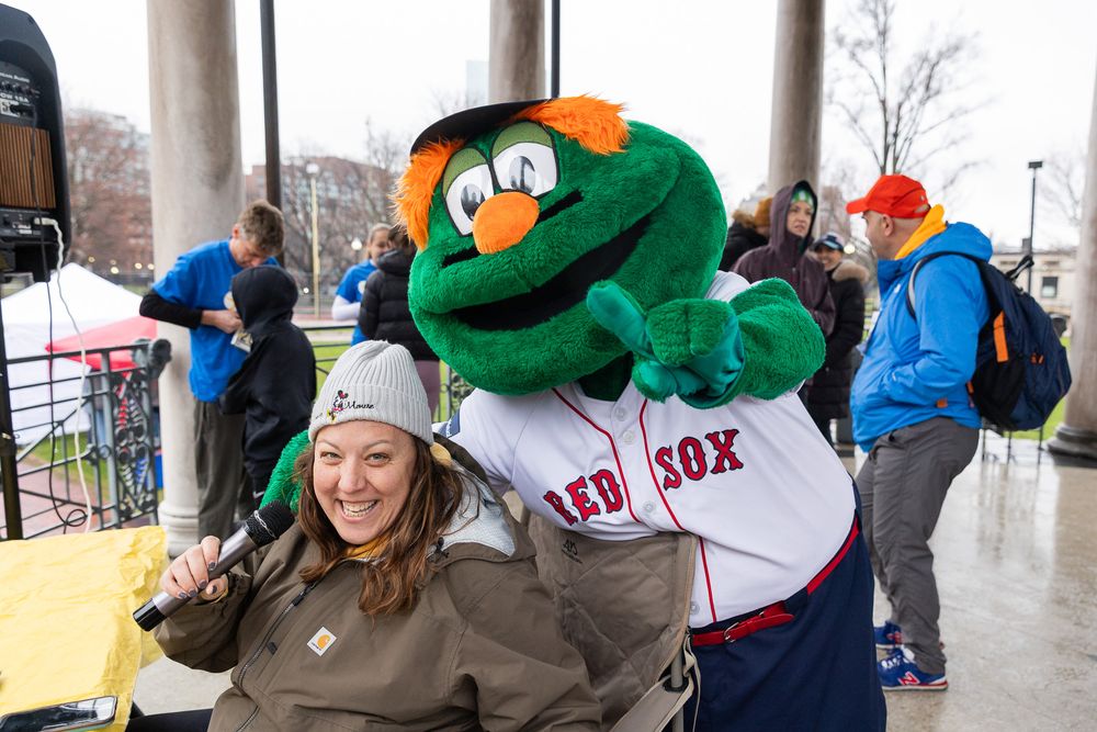 A person in a winter hat laughs with Red Sox mascot Wally the Green Monster at Fenway Park.