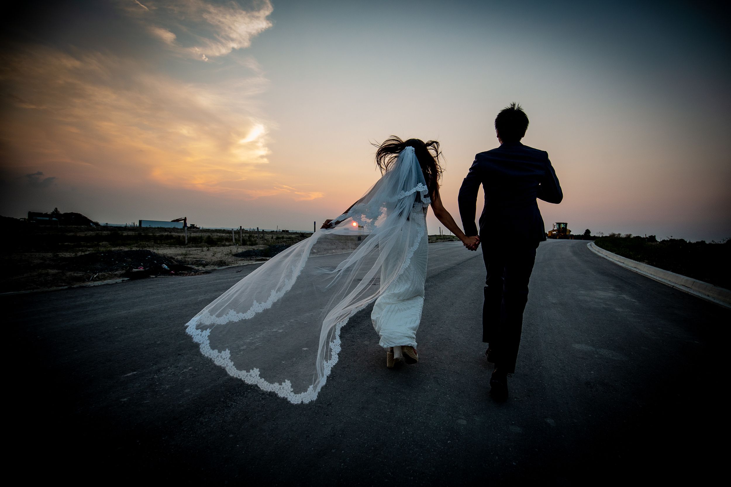 A couple walking on a sunset road with the bride's veil flowing dramatically in the wind.