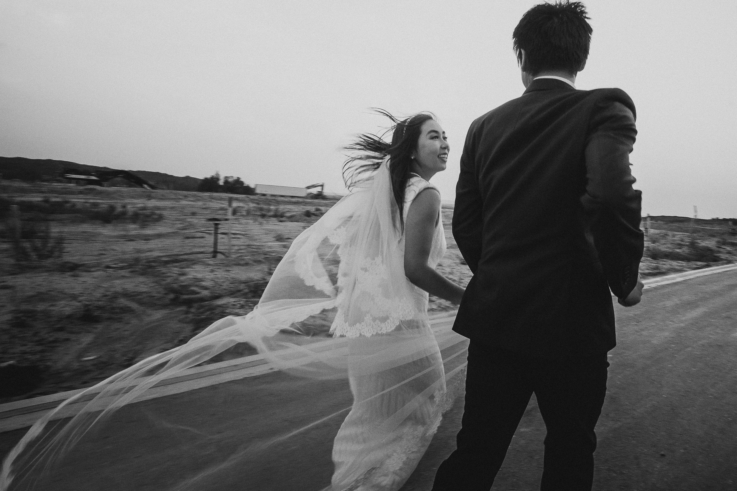 Black and white photo of newlyweds on a rural road with bride's veil billowing in the breeze.