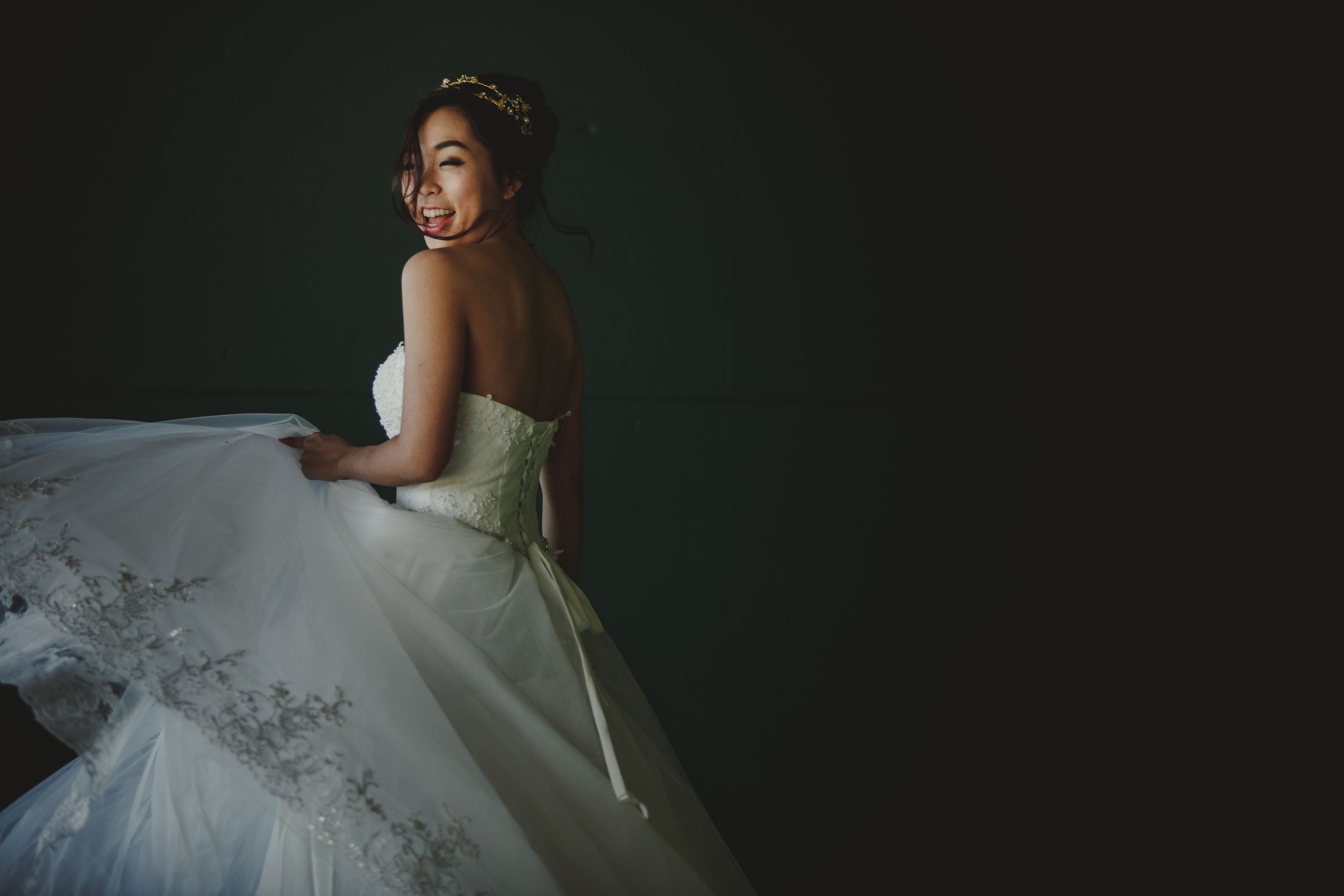 Elegant bride in a strapless ballgown poses against dark backdrop with dramatic lighting and joyful expression.
