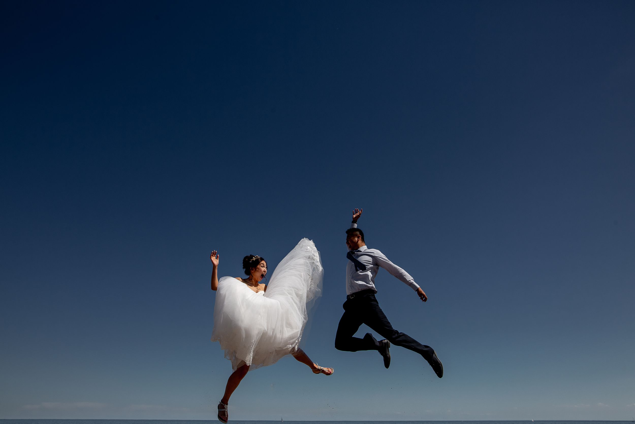 Playful couple leaps joyfully on beach, with bride in flowing white dress against deep blue sky.