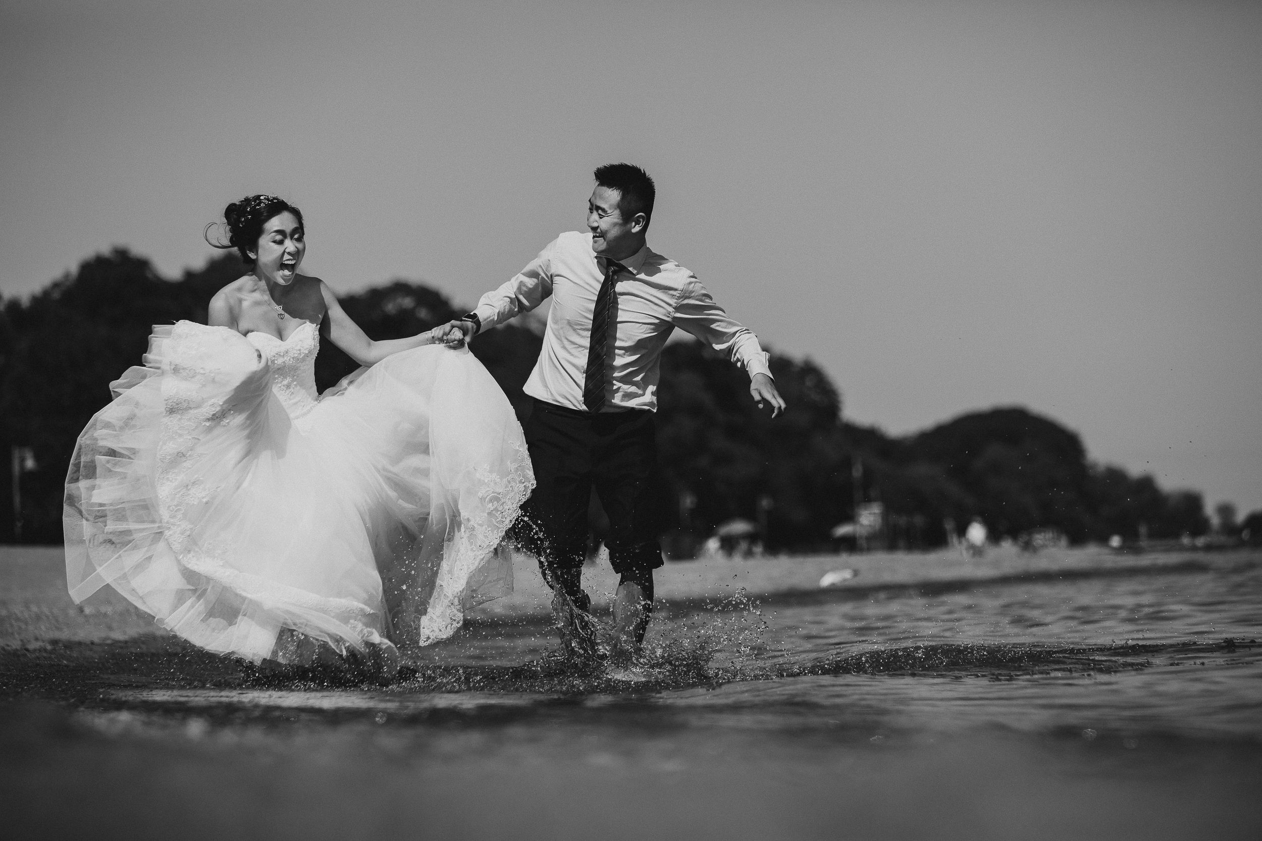 Black and white photo of couple splashing and playing in water wearing wedding attire.